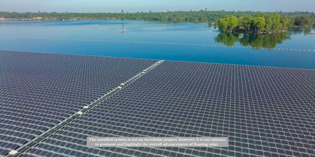 A wide floating solar array on a calm reservoir with trees in the distance, showing solar rafts extending across the water surface as part of a large renewable energy installation.