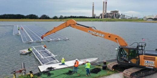 Economic Impact Floating Solar Farms shown through workers assembling floating solar panel platforms on a body of water, with a large excavator positioning the structures and an industrial facility visible in the background.
